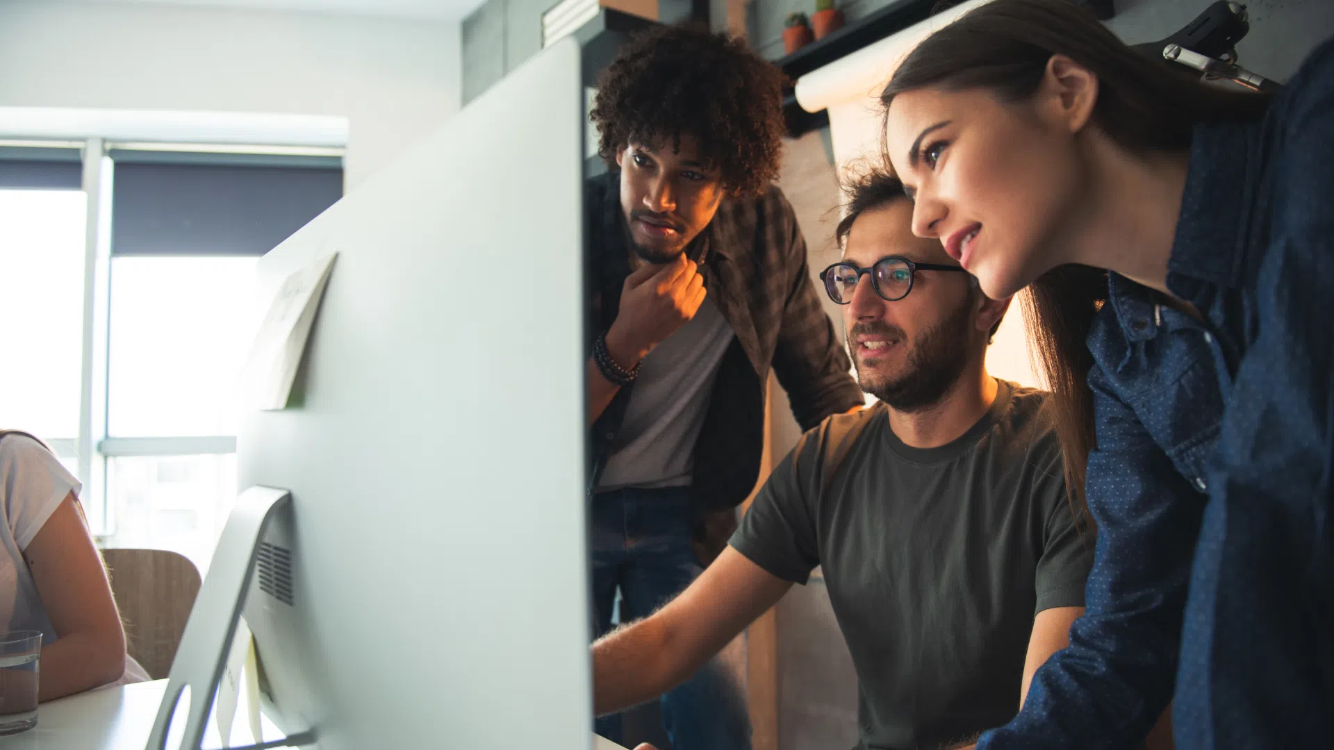 Startup team of diverse young professionals working together around a desktop computer in a modern office environment.