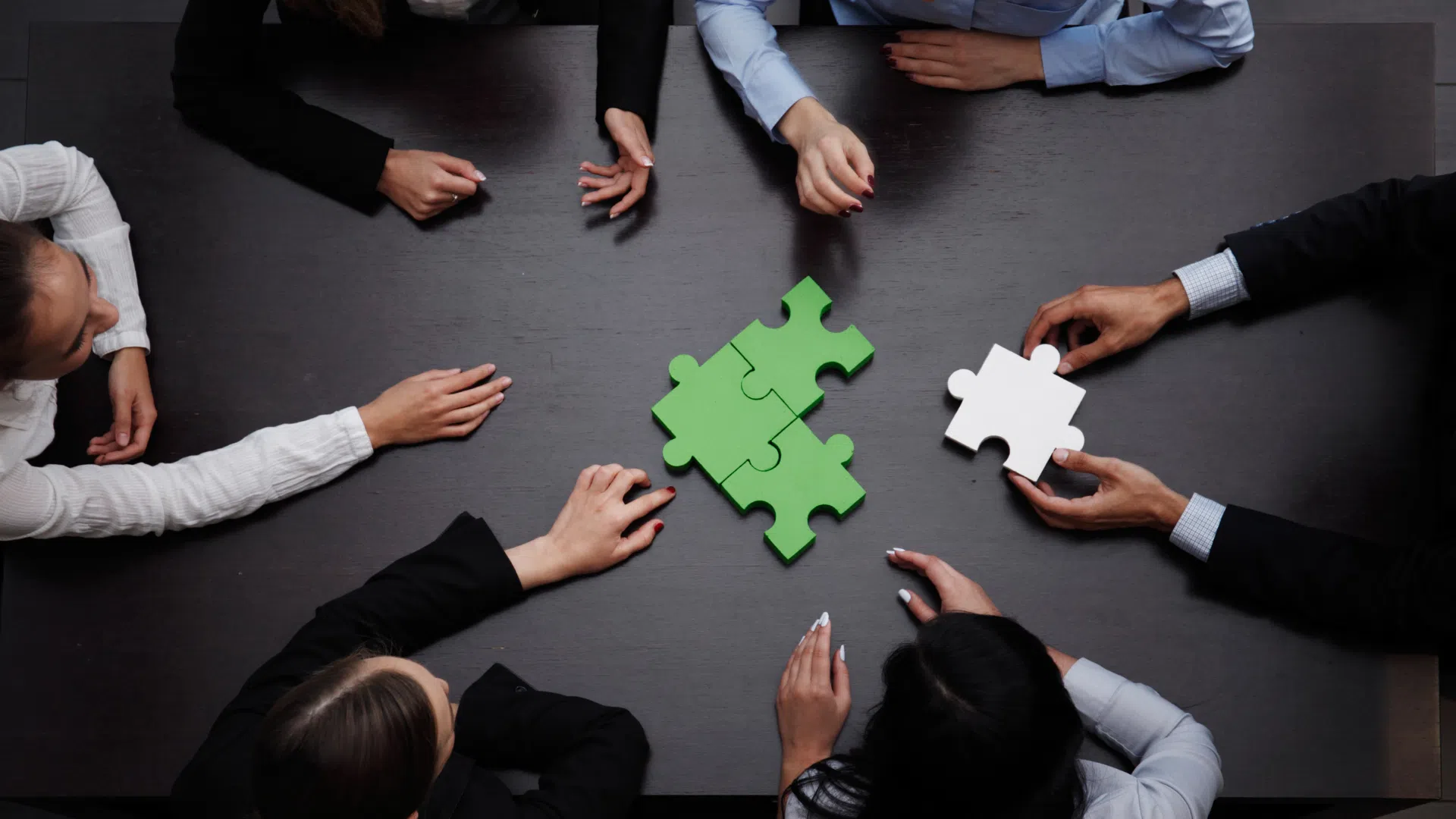 Top-down view of diverse professionals around a table assembling green puzzle pieces, symbolizing team collaboration and strategic problem-solving.